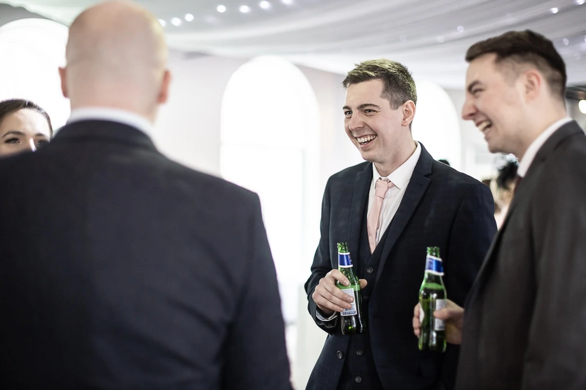 Two men in suits at a social gathering, smiling and holding beer bottles, conversing with a person whose back is to the camera.