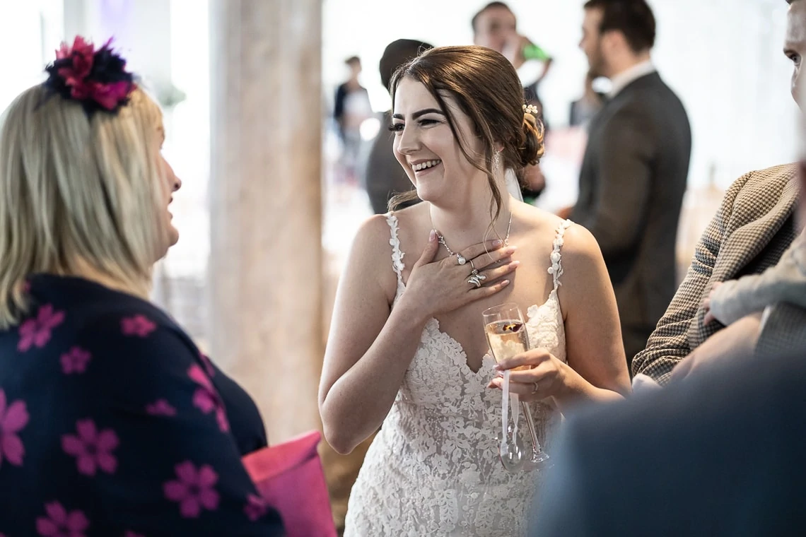 A bride in a lace dress laughs joyfully while holding a champagne glass and talking to a woman with a pink floral headpiece at a wedding reception.