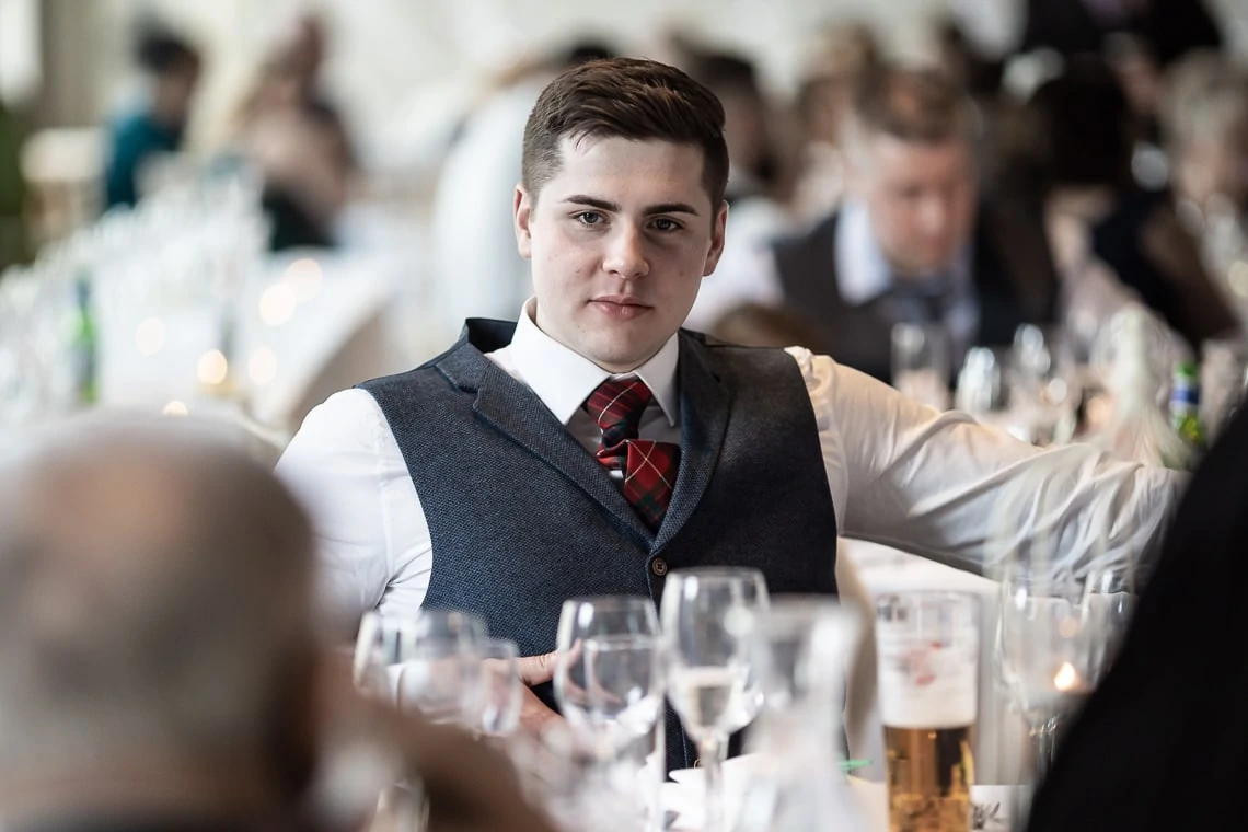 Young man in a vest and tartan tie attending a formal event, pausing as he interacts with guests at a dining table.
