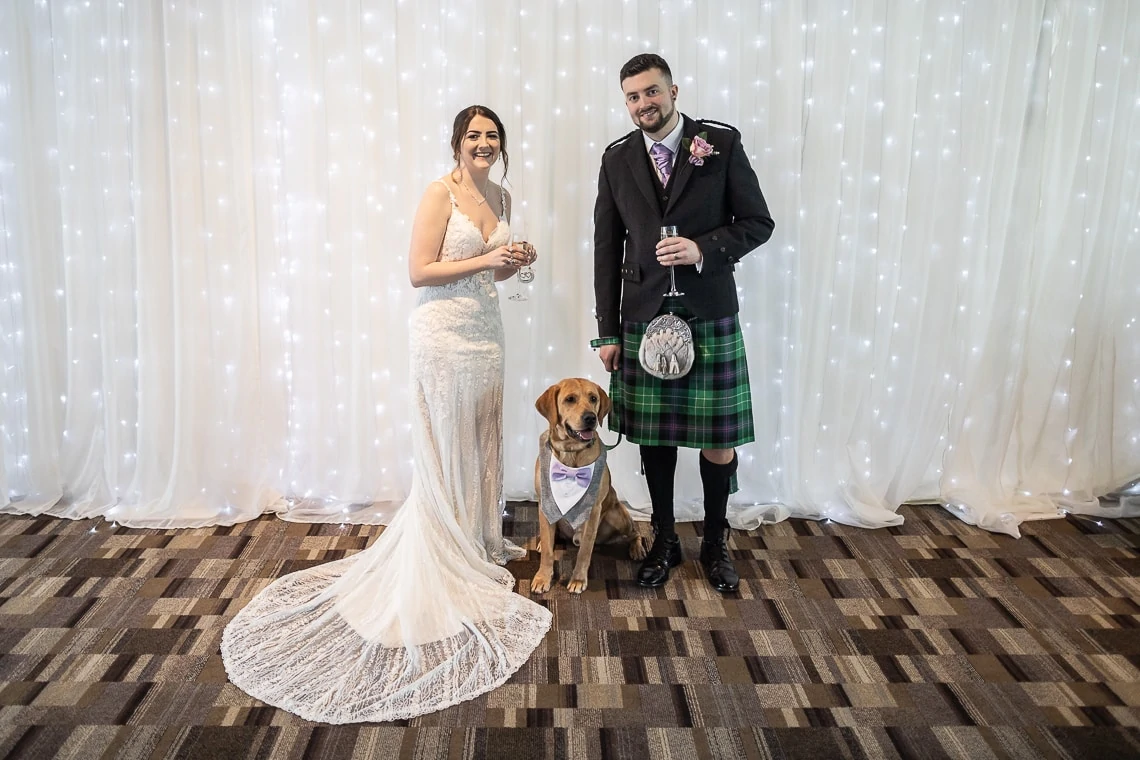 A bride in a beaded gown and a groom in a kilt smiling with a golden retriever, standing in front of a white curtain backdrop.