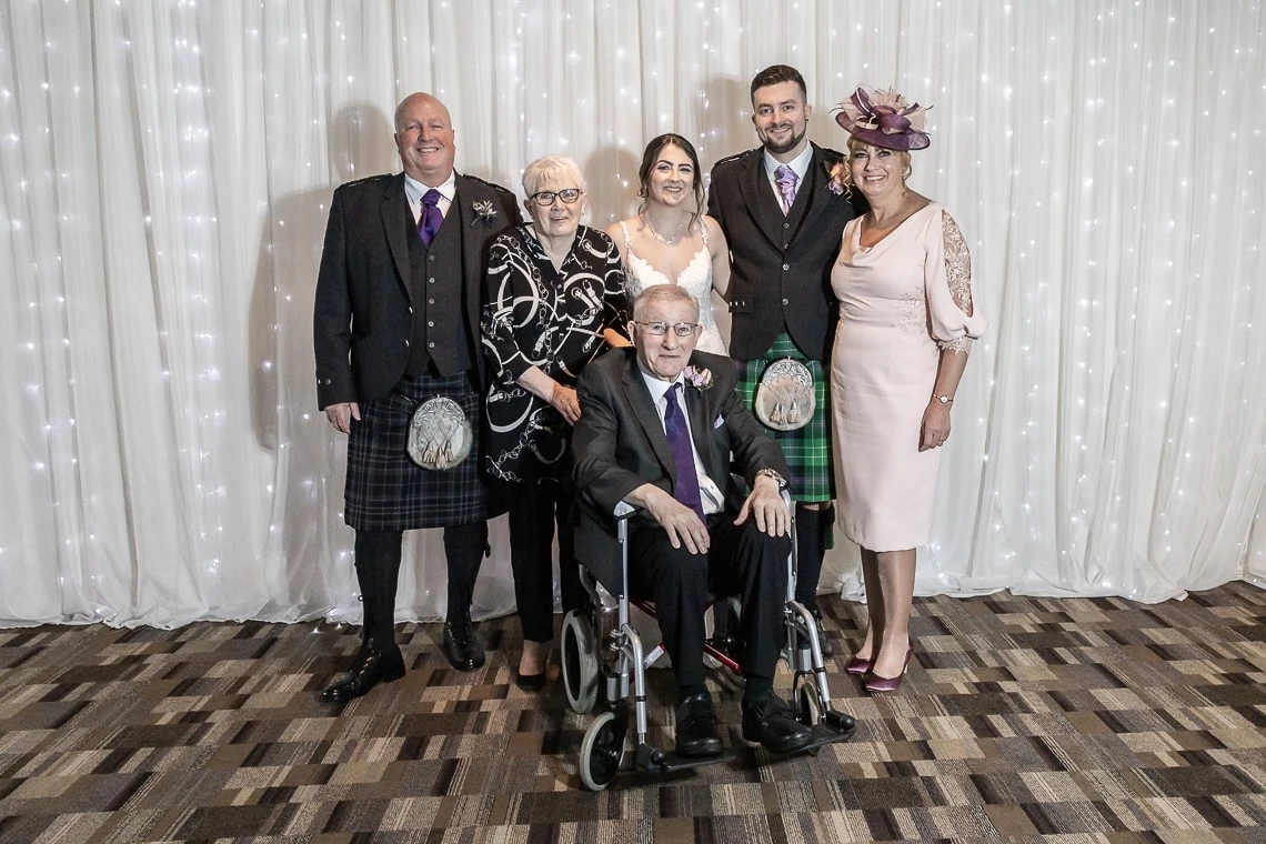 A family of six, with two men in kilts and a seated elderly man, posing for a photo at a formal event with a draped white backdrop.