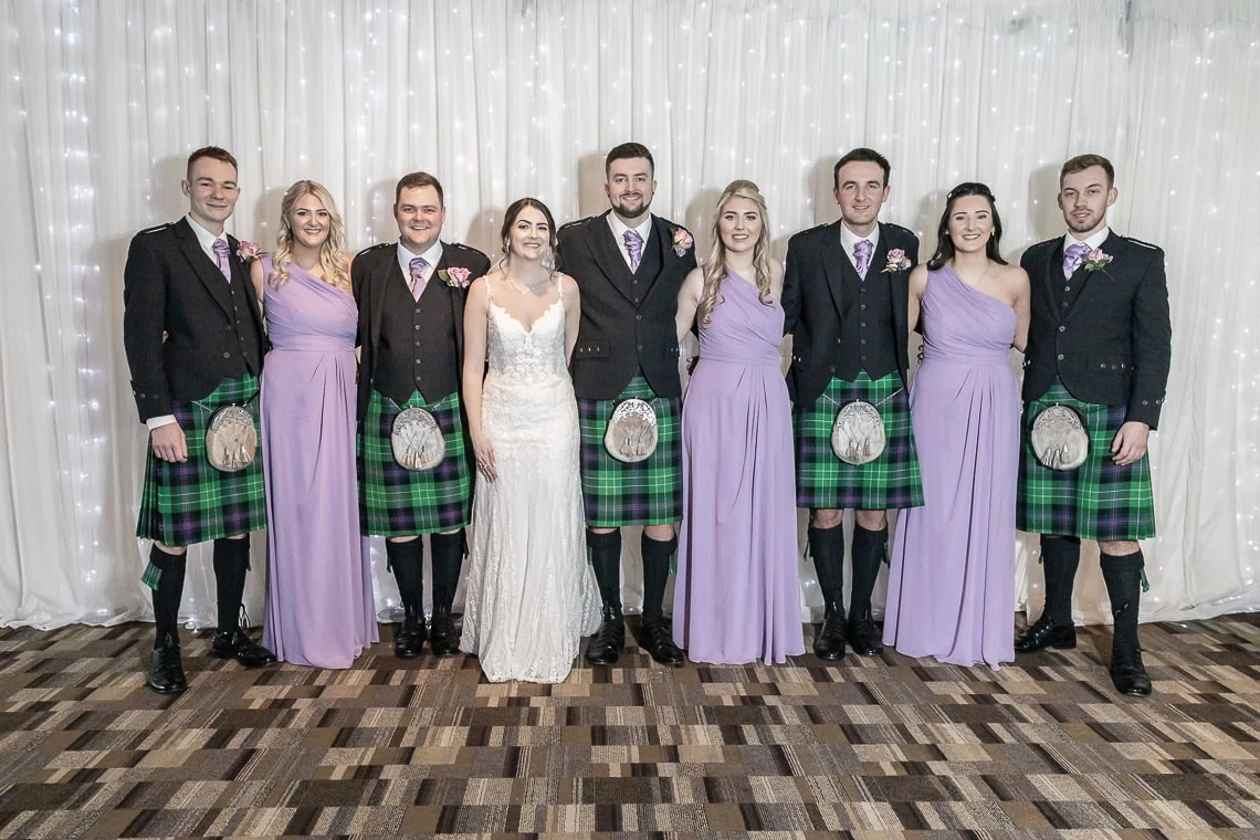 A bridal group posing for a photo; the men wear kilts and the women are in lavender dresses, with the bride in a white gown, all standing in front of a white curtain.