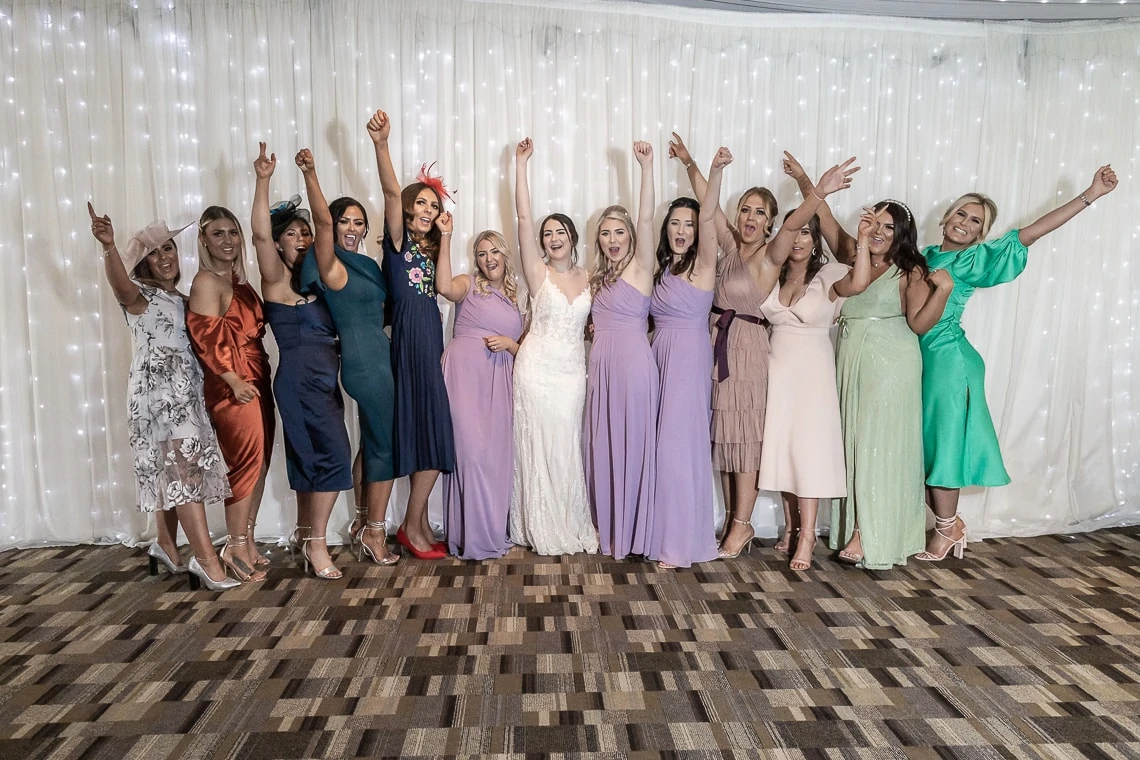 Group of women in formal dresses, celebrating and raising their arms in joy at a wedding event, with a white curtain backdrop.