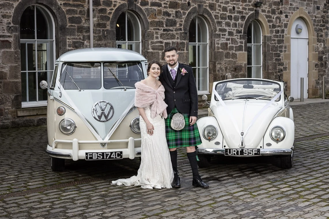 A bride in a lace dress and a groom in a kilt stand between a vintage vw bus and a beetle on a cobblestone courtyard.