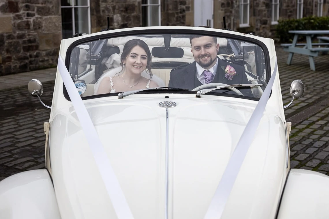 A bride and groom smiling in a white vintage car decorated with ribbons, parked in front of an old stone building.