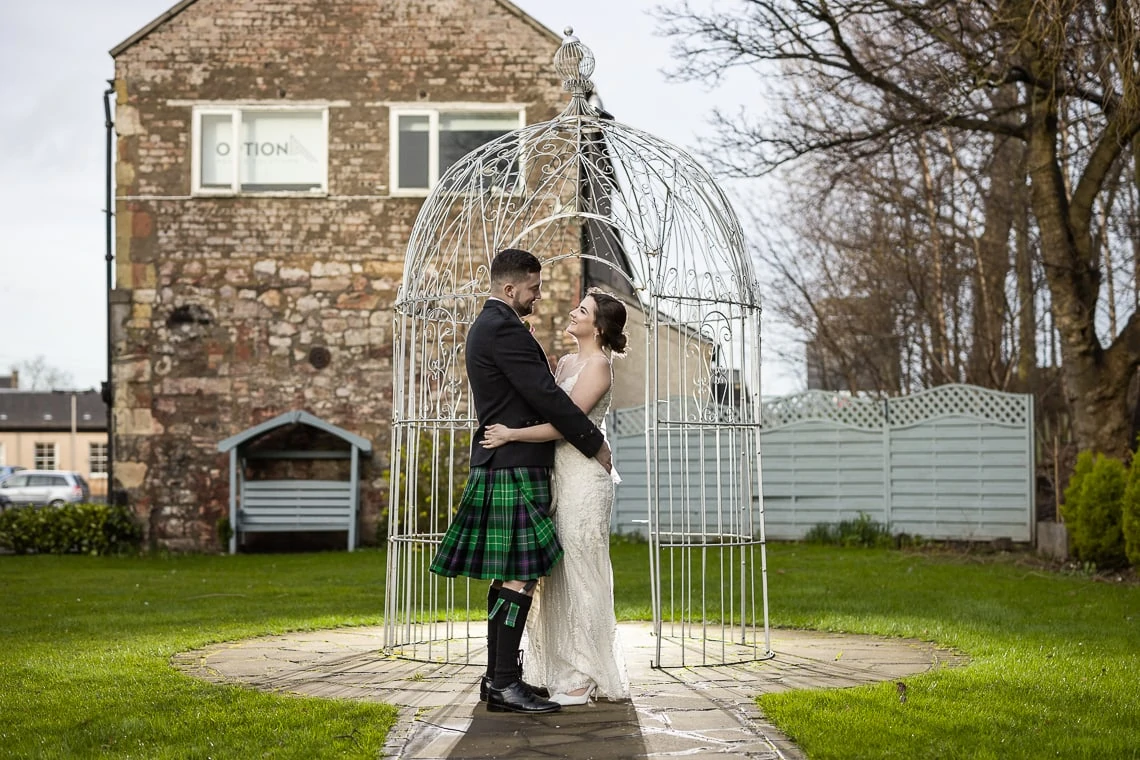 A newlywed couple embracing inside a birdcage-shaped structure in a garden, with the groom wearing a tartan kilt and the bride in a white dress.