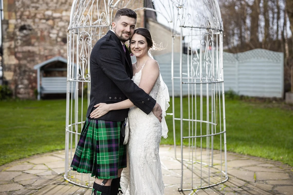 A bride in a white dress and a groom in a kilt embracing in front of a decorative birdcage structure in a garden.