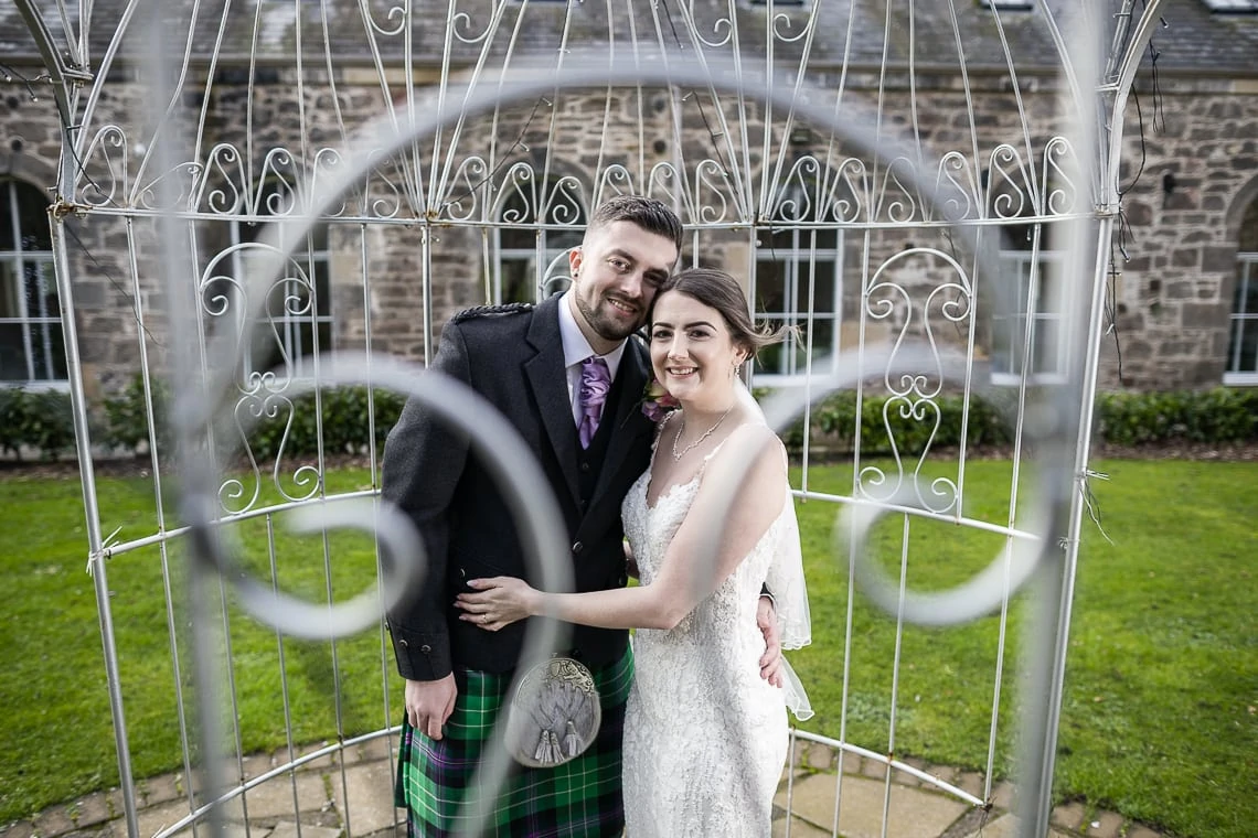 A bride and groom smiling through a decorative metal heart outside a stone building, the groom in a tartan kilt and the bride in a white dress.