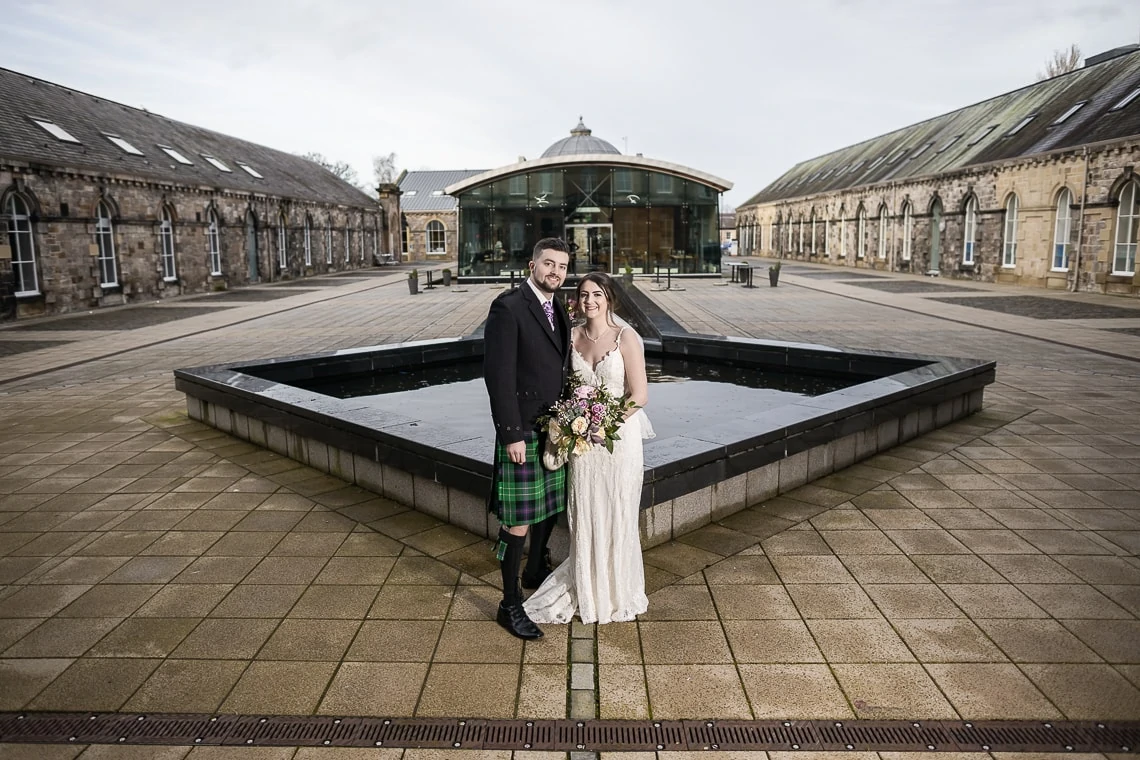 A bride and groom, the groom in a kilt, posing by an octagonal fountain in a spacious courtyard with classical buildings in the background.