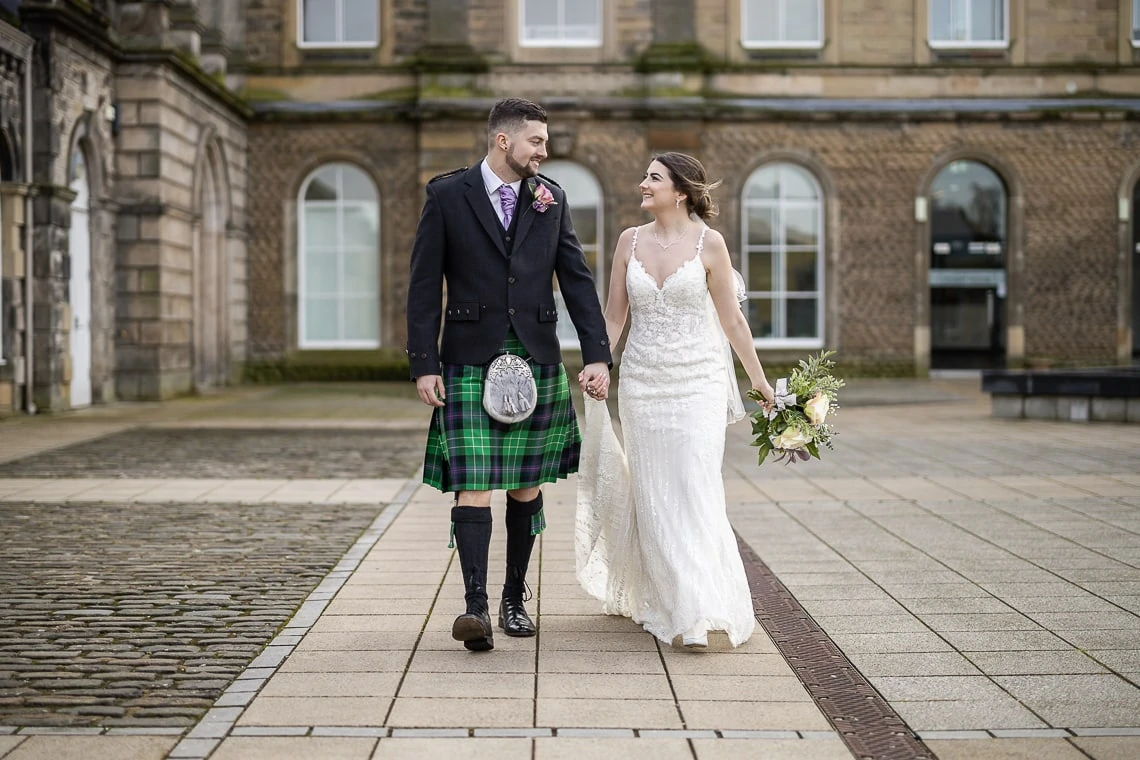 A bride and groom walking hand in hand, the groom in a kilt and the bride in a white dress, in front of a classic building.