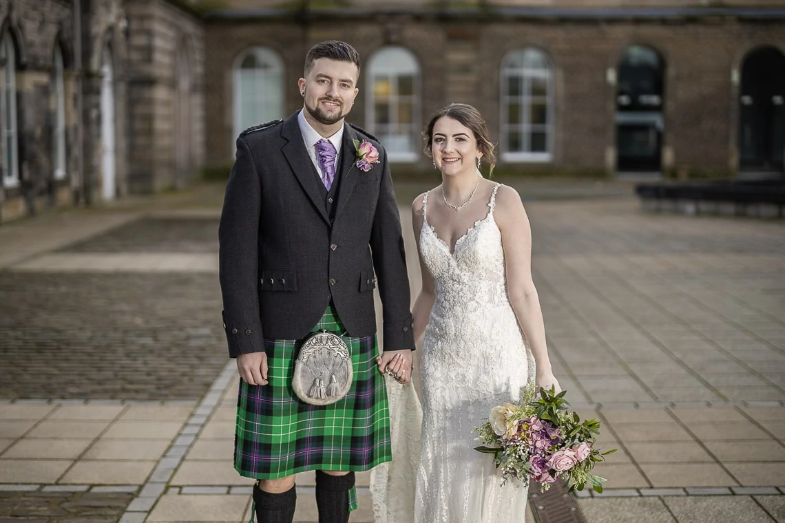 A bride and groom smiling in formal wedding attire; the groom is wearing a kilt with a tartan pattern and sporran, the bride a sleeveless lace gown, holding a bouquet, in front of a historic building.
