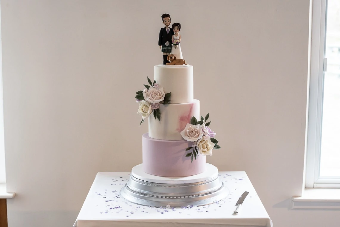 A three-tiered wedding cake with purple and white layers, decorated with fresh roses and a topper featuring a couple with a dog.
