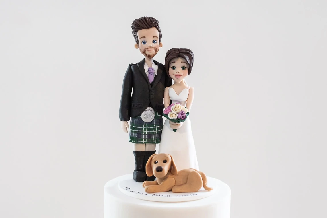 A wedding cake topper featuring a clay figurine of a bride and groom with a small dog, set on a white cake base.