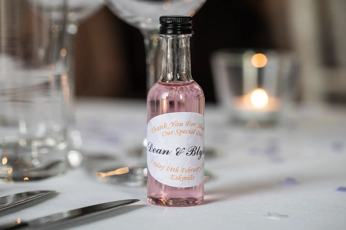 A small bottle of pink liquid with a custom label "thank you for sharing our special day" on a table set for a celebration, with soft background lighting.