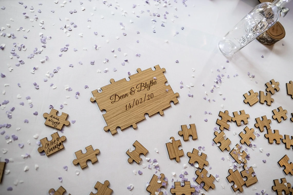 A wooden puzzle piece engraved with "dave & blythe 10/07/20" surrounded by scattered puzzle pieces and lavender petals, with a champagne glass tipped over.