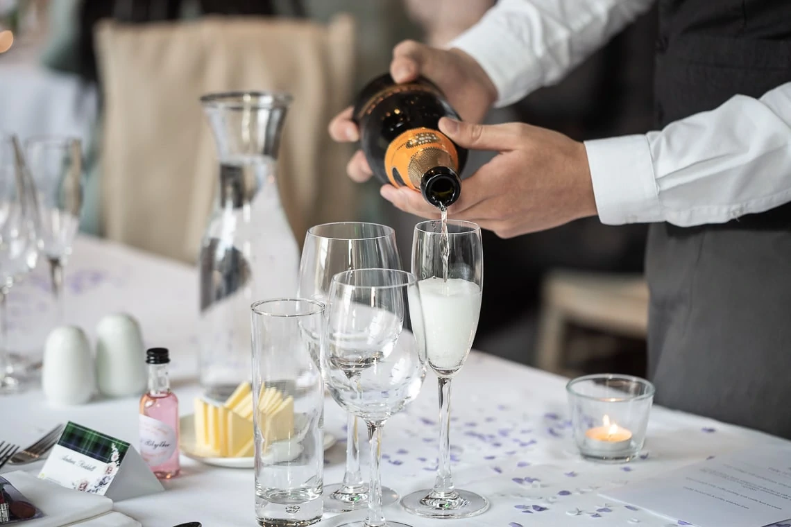 A waiter pours champagne into a flute at a neatly arranged table with empty glasses and a lit candle, in a restaurant setting.