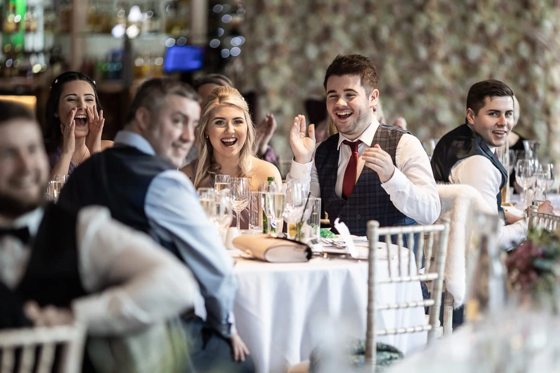 Group of people at a wedding reception table laughing and interacting, with a focus on a young man smiling and clapping.