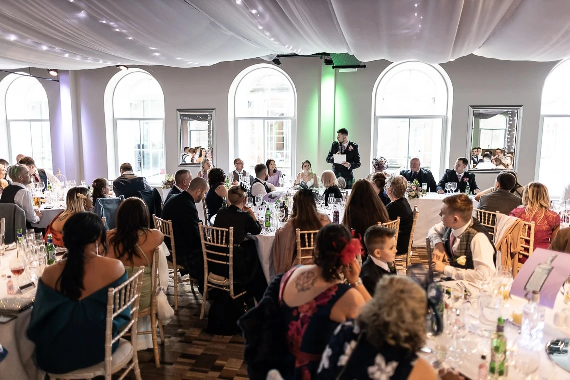 Elegant wedding reception with guests seated at round tables listening to a man giving a speech.
