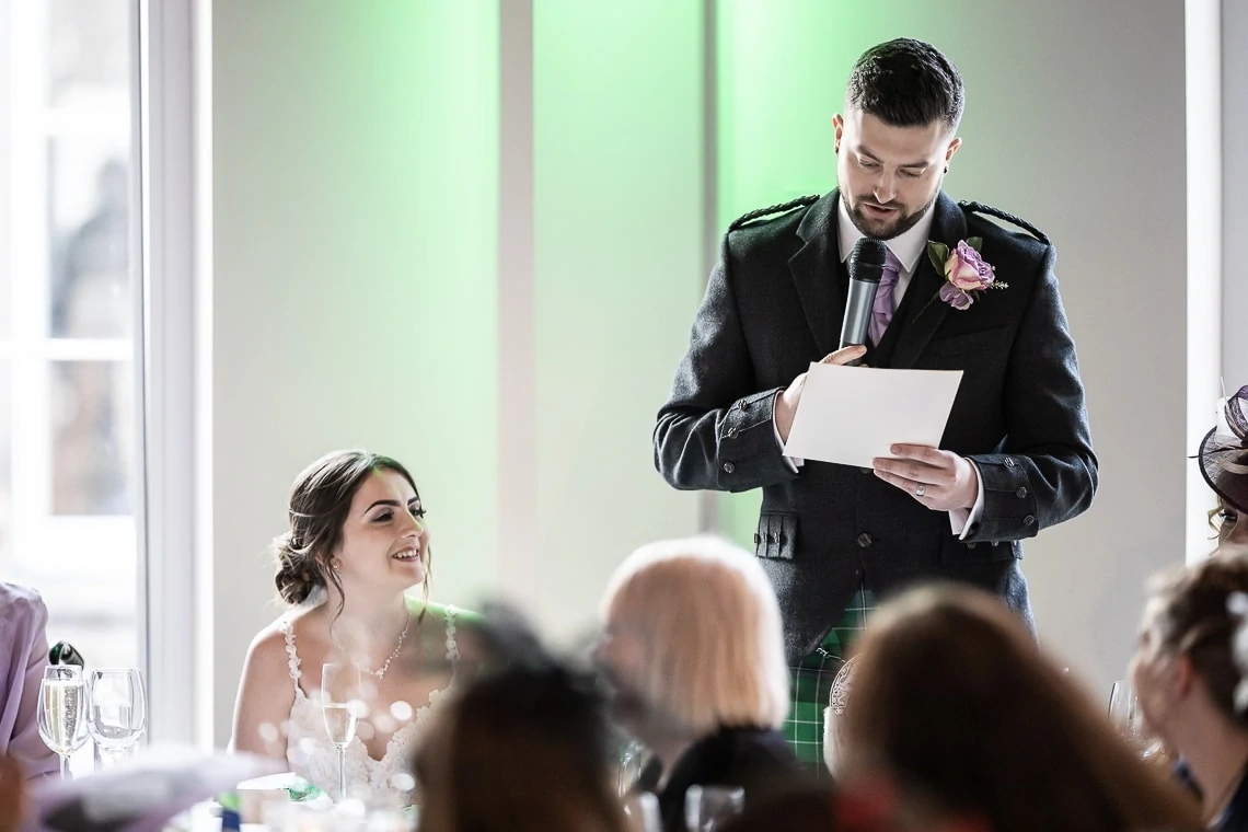 A man in a formal suit reads from a paper at a wedding reception, with a smiling woman in the background by a window.