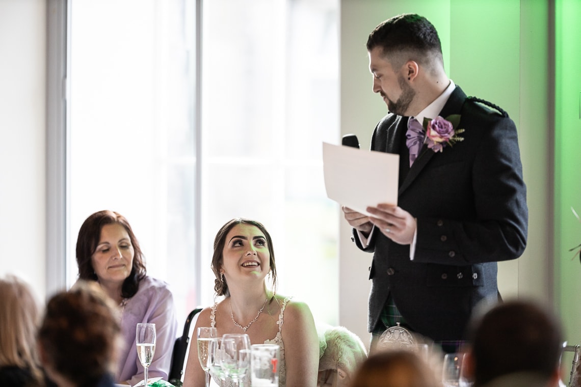 A man in a suit and boutonniere reads from a paper during a wedding reception, while a smiling woman in a white dress looks up at him.