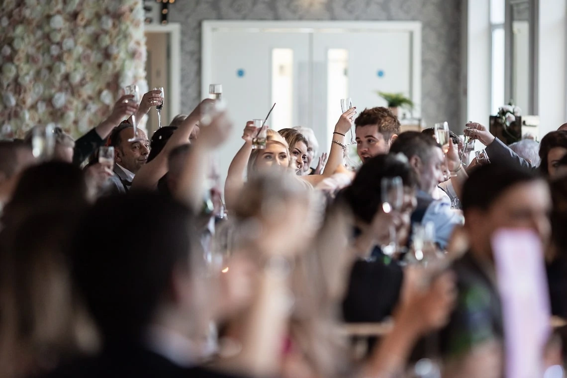 Guests raising glasses for a toast at a wedding reception, focus on a couple among the crowd.