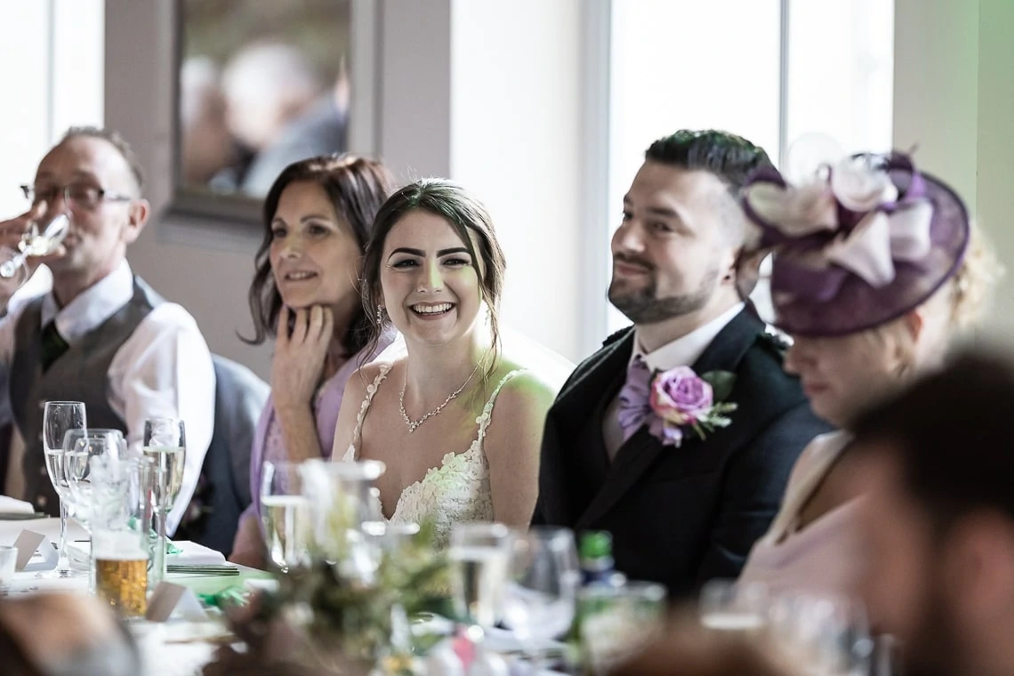 A bride and groom smiling at a wedding reception table with guests around them, one wearing a purple hat.