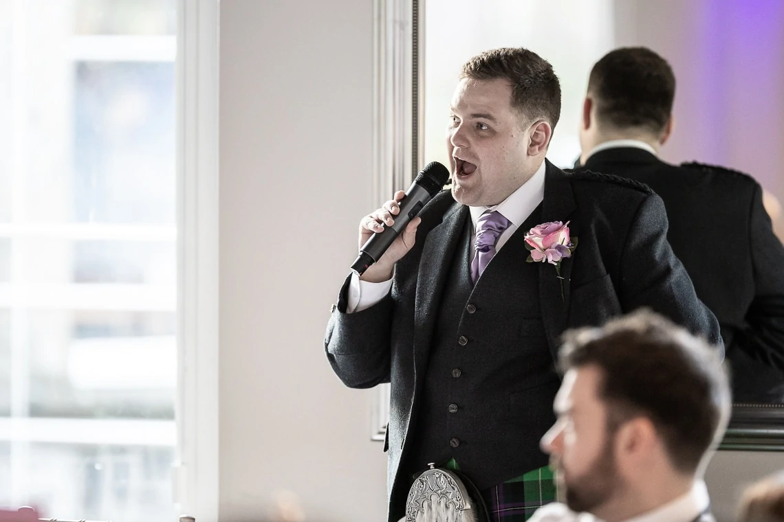 Man in traditional scottish attire speaking into a microphone at an event, with a focus on his expressive face and detailed outfit.