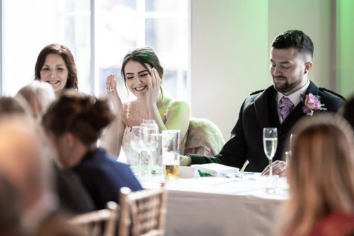 A bride in a green dress laughs joyfully at a wedding reception table with other guests, including a groom in a tartan kilt.