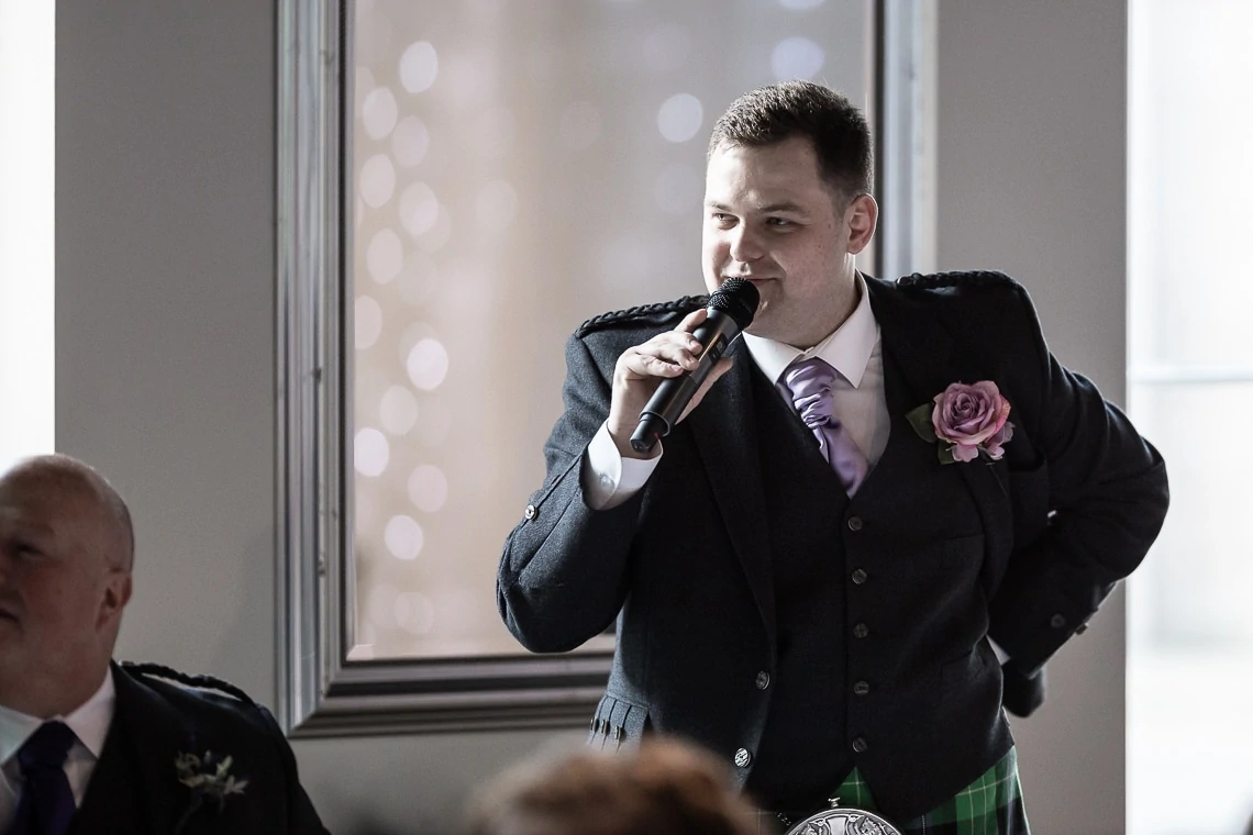 A man in a kilt and suit jacket speaks into a microphone at an event, with a blurred background and a window beside him.