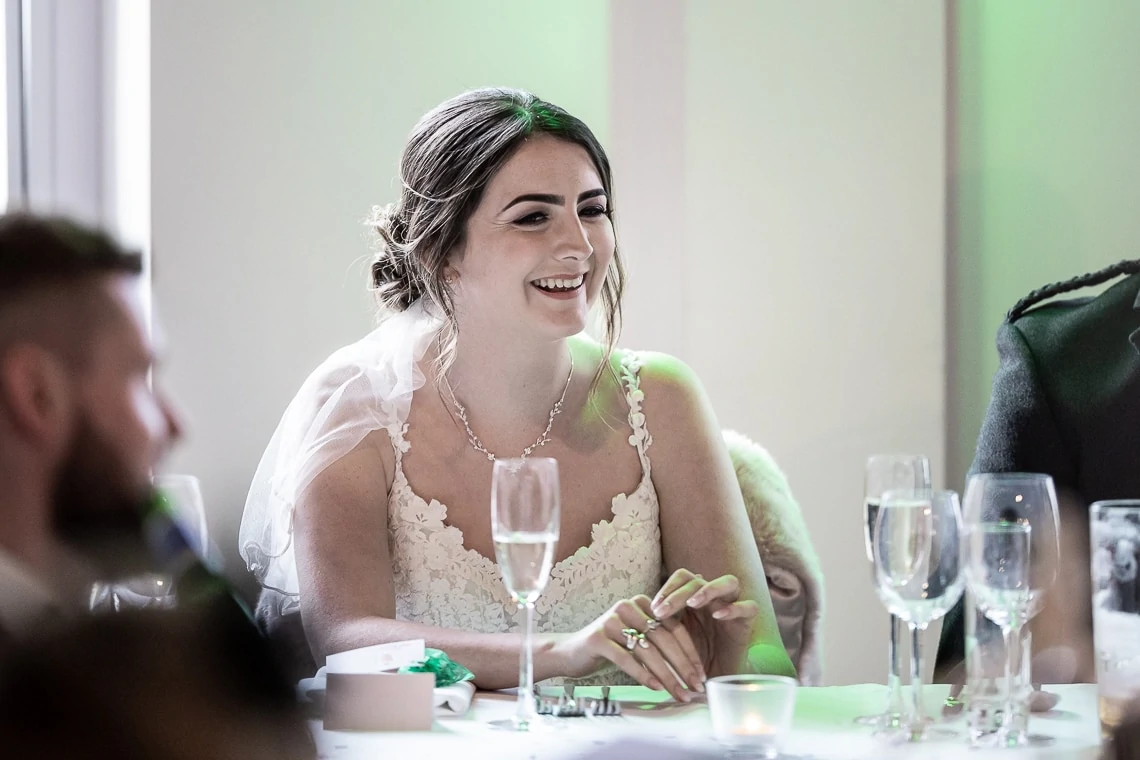 A bride in a lace dress smiling at a wedding reception table, surrounded by guests and glasses.