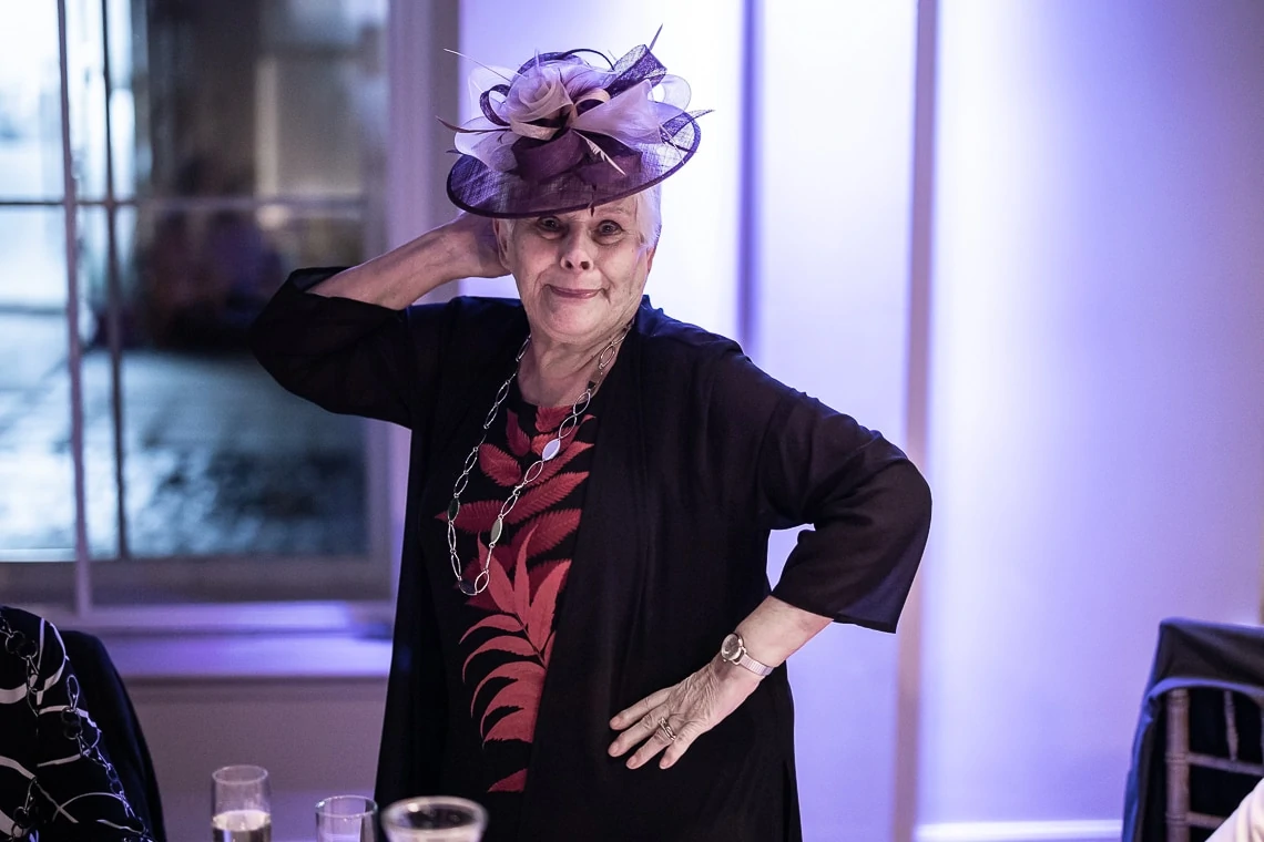 Elderly woman wearing a purple hat and black outfit posing with her hand on her hip at a social event.