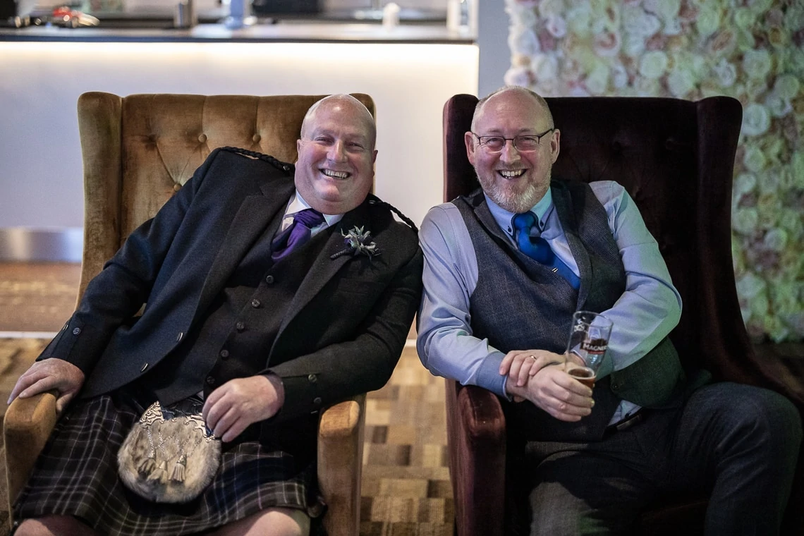 Two smiling men in traditional scottish attire, including kilts, seated on an ornate couch with drinks in hand.