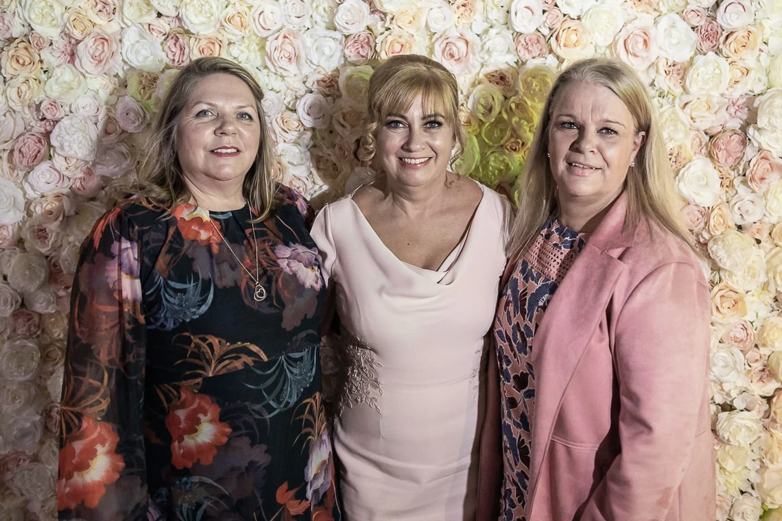 Three smiling women pose in front of a floral backdrop, the middle woman in a pink dress flanked by two others in patterned outfits.