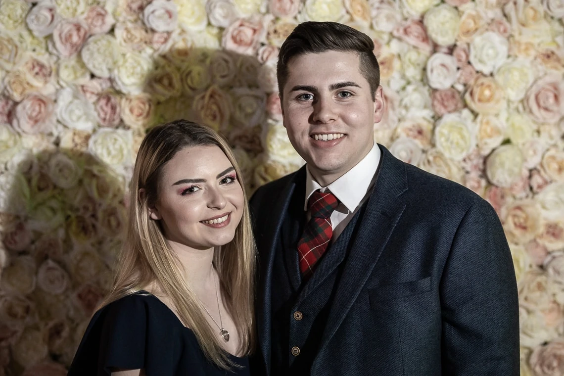 A young couple smiling at the camera with a floral backdrop, the woman in a navy dress and the man in a grey suit and plaid tie.