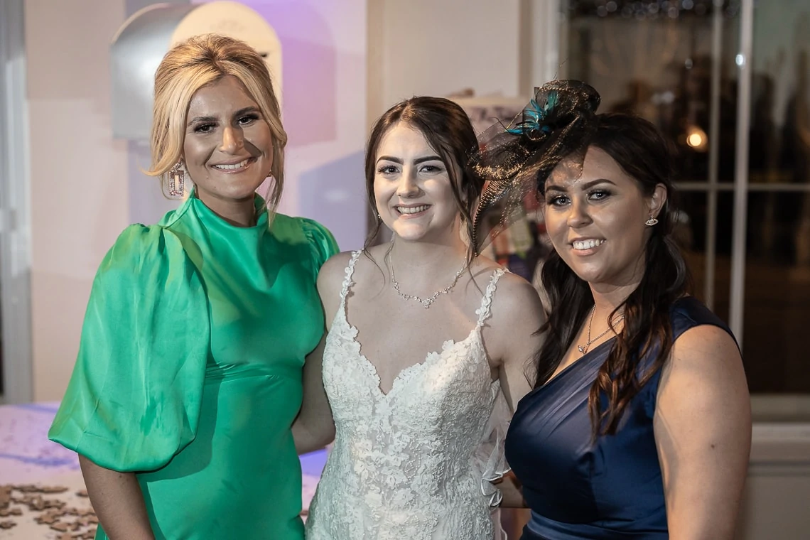 Three women smiling at a formal event, with one in a wedding dress flanked by two others in elegant green and blue dresses.