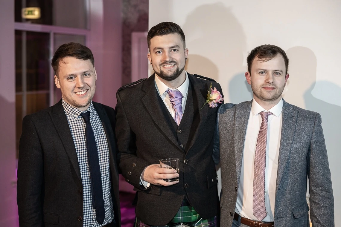 Three men at a formal event, one in a kilt and tweed jacket holding a drink, flanked by two others in suits, smiling at the camera.