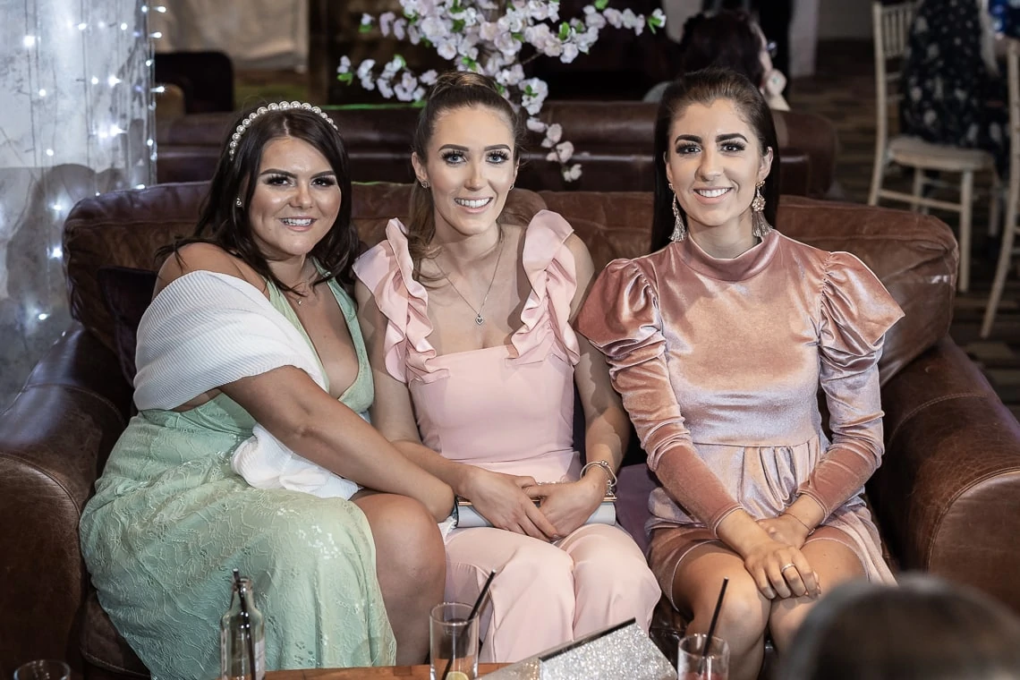 Three women smiling, seated together at a social event, wearing formal dresses in pastel shades.