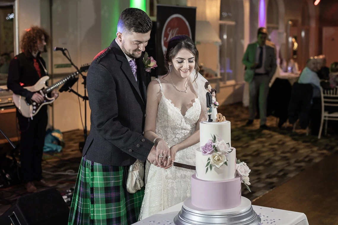 A bride and groom cut a multi-tiered wedding cake together at their reception, the groom wearing a kilt and the bride in a white gown.