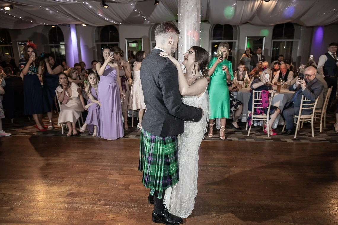 A bride and groom share a dance at their wedding reception, with guests surrounding them in a brightly lit hall. the groom wears a tartan kilt.