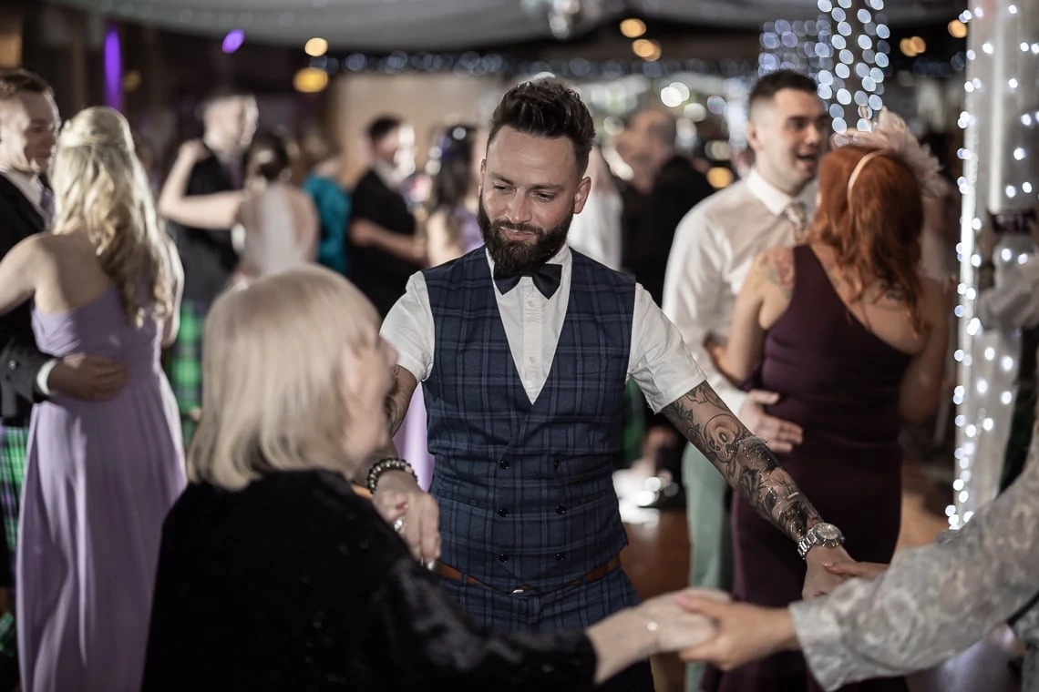 A man in a vest and bow tie dances joyously with guests at a festive indoor party, with other attendees visible in the background.