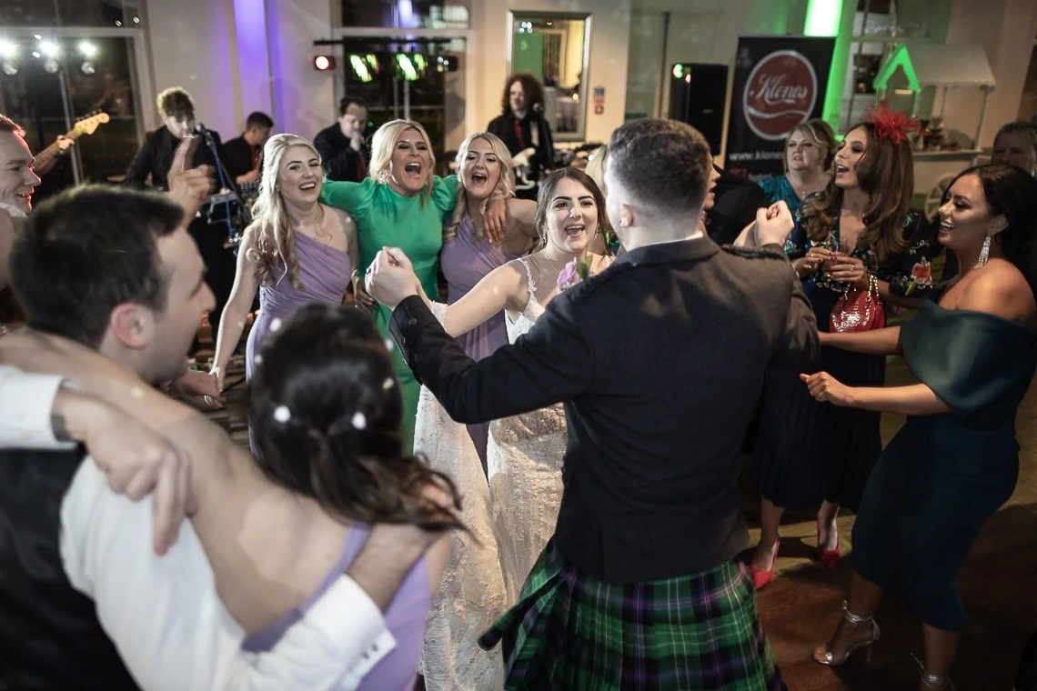 A bride, groom in a kilt, and guests laugh and dance joyously at a wedding reception with a live band in the background.