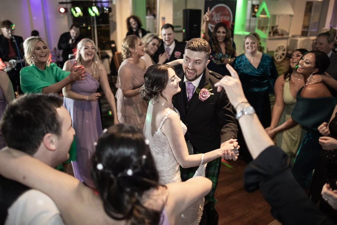 A bride and groom dance joyously among a crowd of guests at their wedding reception, with smiling attendees clapping around them.