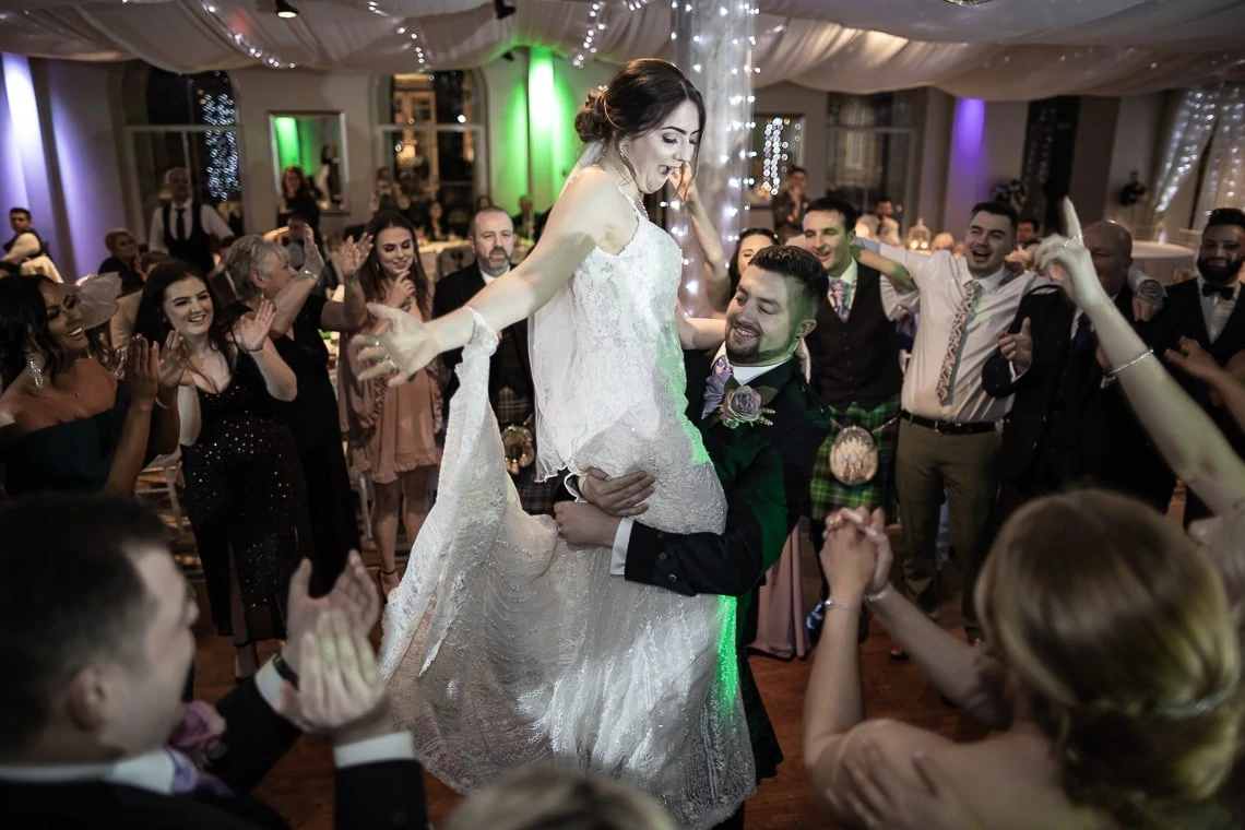 A groom lifts his bride during their wedding dance, surrounded by applauding guests in a festively decorated hall.