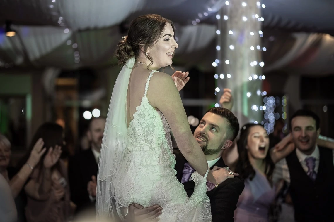 Bride being lifted by the groom during a wedding reception dance, surrounded by cheering guests under twinkling lights.