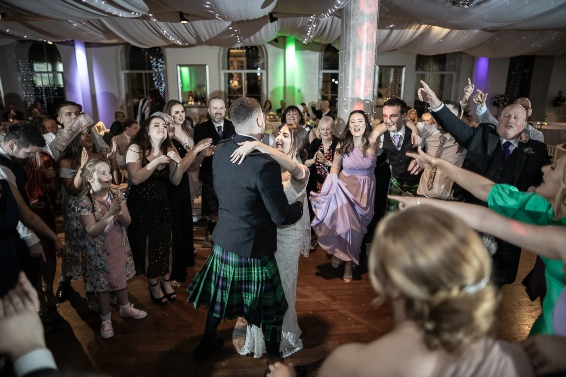 A joyful wedding reception scene with guests clapping around a dancing couple, the groom in a kilt, inside a festively lit hall.