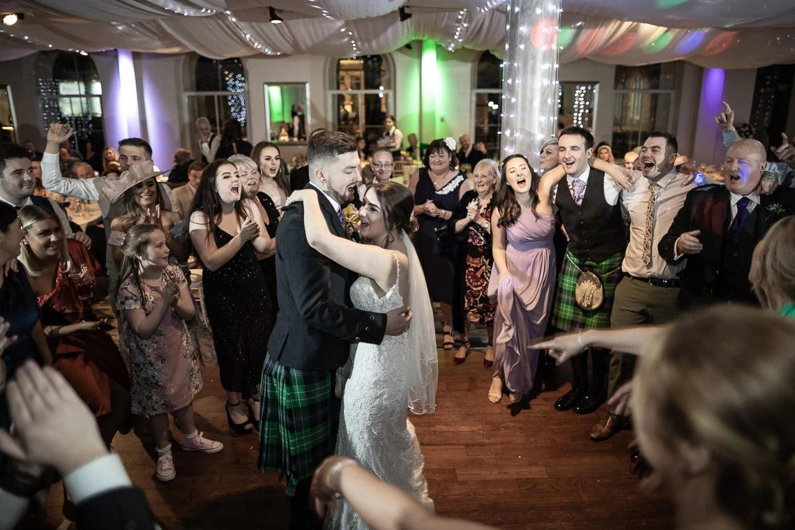 A bride and groom dancing joyfully at their wedding reception, surrounded by cheering guests in a festively decorated hall.