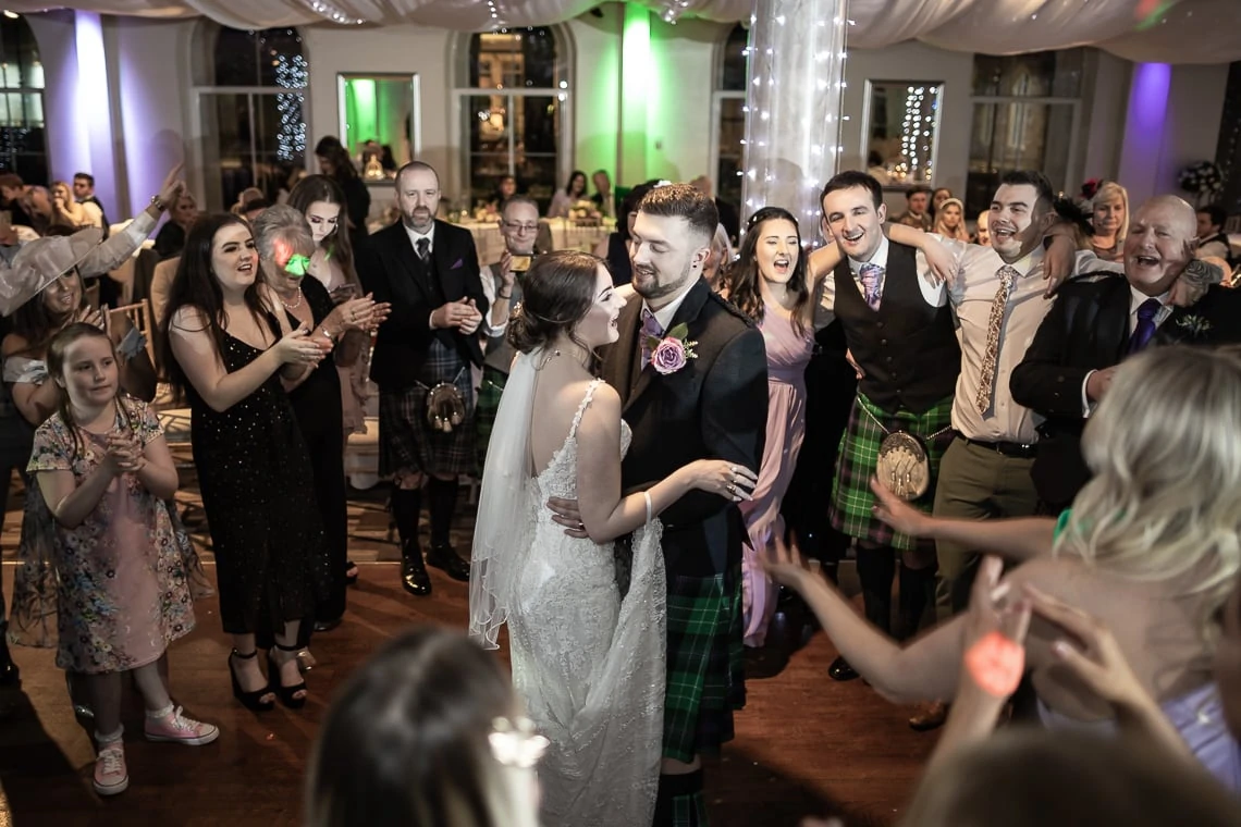 A bride and groom walk through a crowd of clapping guests, many in kilts, during a lively reception in an elegantly decorated hall.