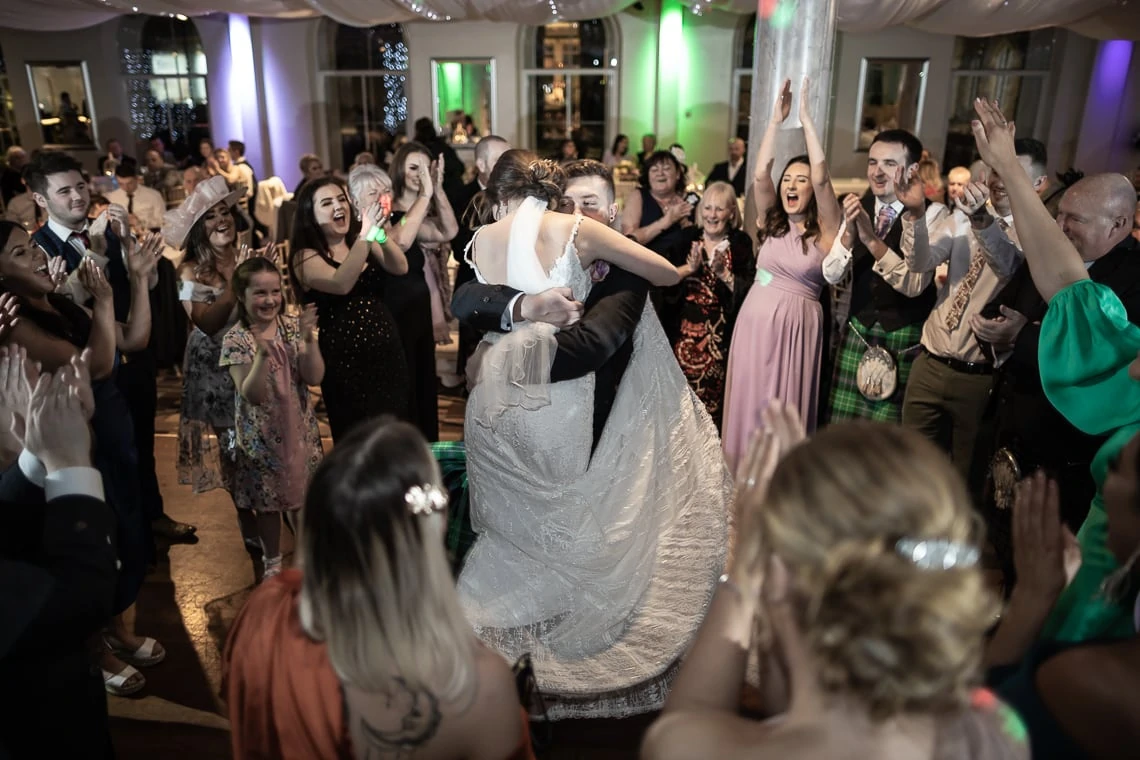 A bride and groom share a kiss while dancing, surrounded by cheering guests at a wedding reception.