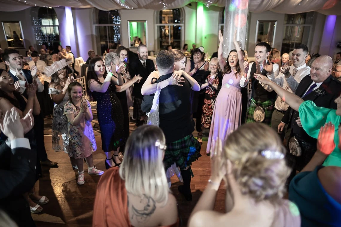 A couple dances joyously at their wedding reception, surrounded by cheering guests in a festively decorated hall.