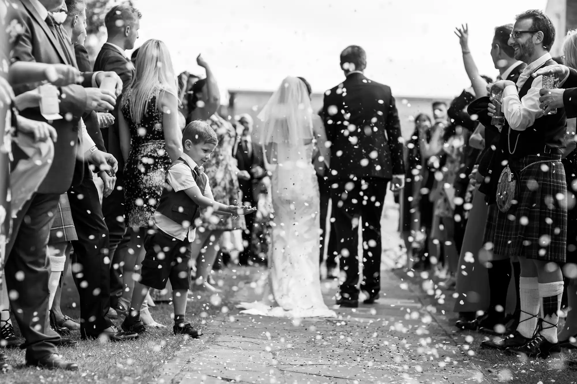 Black and white image of a wedding celebration with guests throwing confetti as a bride and groom walk down an outdoor aisle.